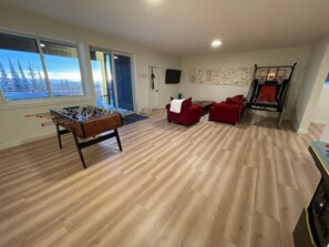 A living room with a foosball table, red sofas, a basketball hoop, and a large window with a view of snow-covered trees. - Create unforgettable memories in a home designed for comfort, and adventure. (Vernon)