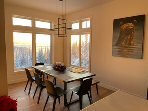 A dining area with a table set for four, large windows with a view of snow-covered trees, and a bear painting on the wall. - Create unforgettable memories in a home designed for comfort, and adventure. (Vernon)