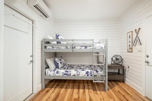 Ein Schlafraum mit Etagenbetten, weißen Wänden und Holzboden. Ein Ventilator steht im unteren Bett, und auf der linken Seite befindet sich eine Tür.