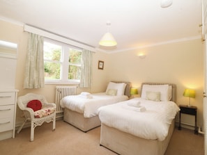 Ein Hotelzimmer mit zwei Einzelbetten, einem weißen Schminktisch, einem Korbstuhl, einer Heizung und einem Fenster mit Vorhängen.