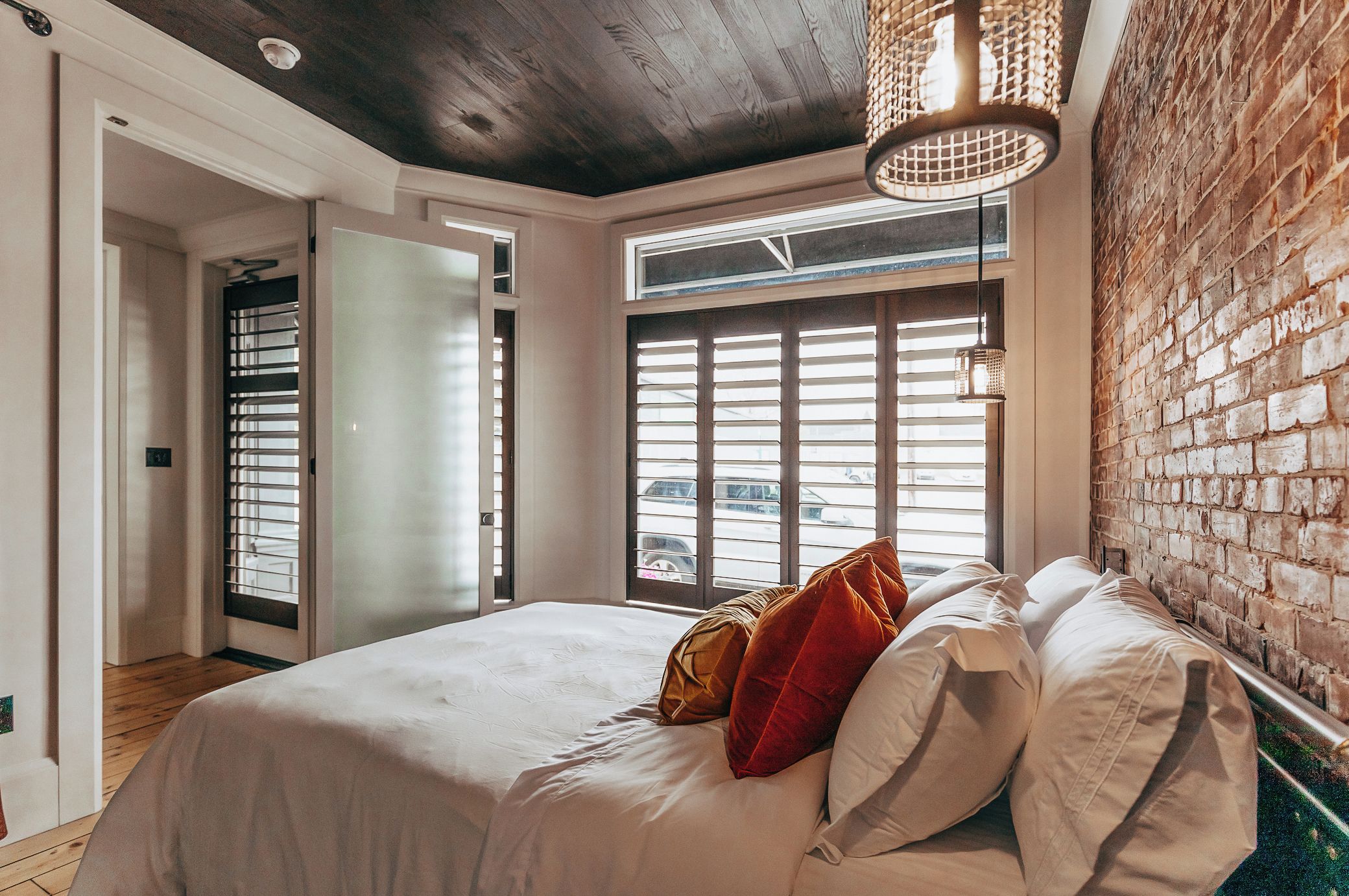 A bedroom with a bed, a window with blinds, a brick wall, and a ceiling light fixture.