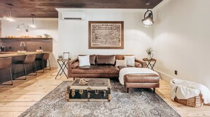 A modern living room with a brown leather sofa, a patterned rug, a vintage trunk, and a framed picture on the wall.