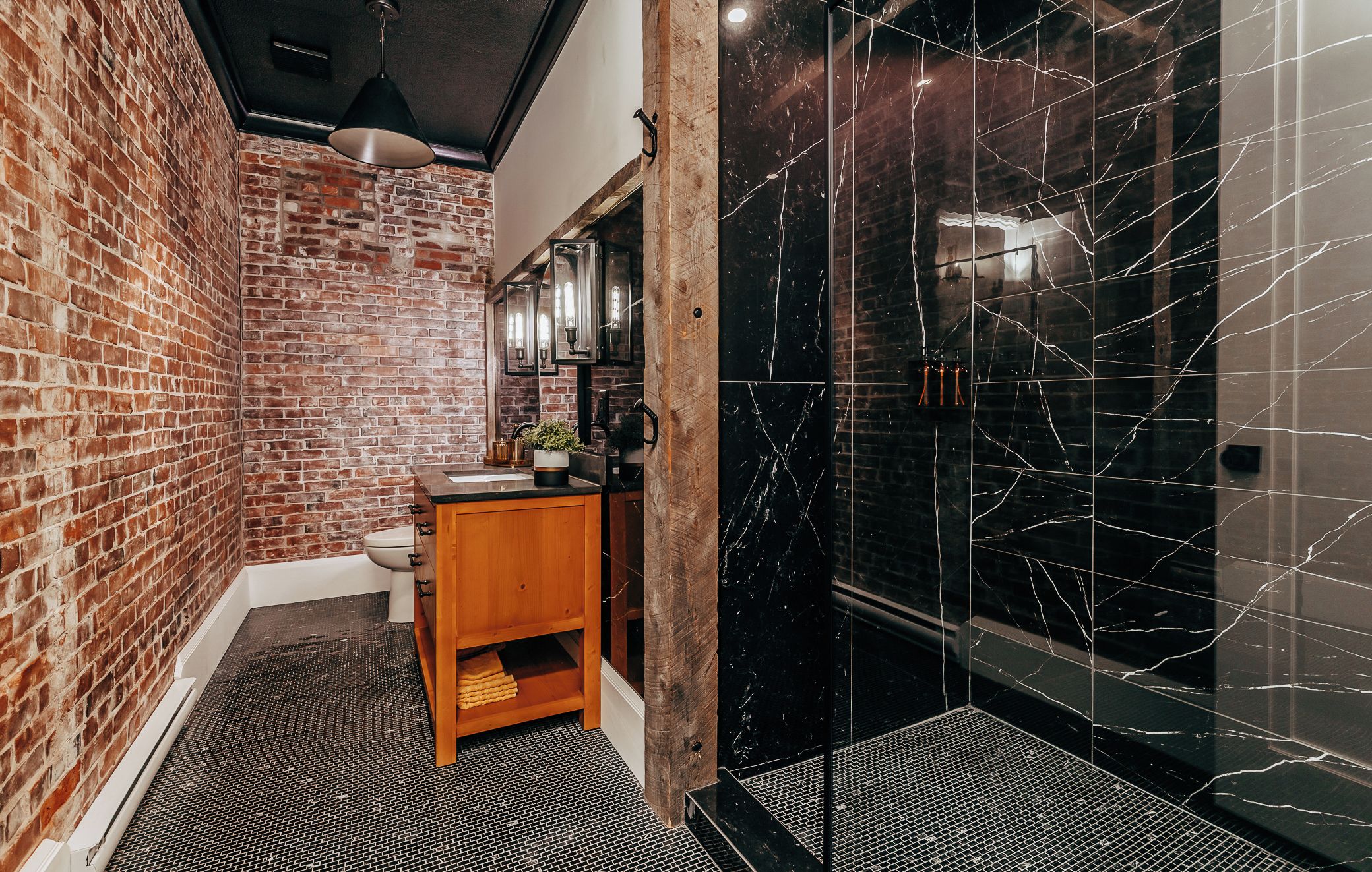 A bathroom with a brick wall, a wooden vanity, a black marble shower enclosure, and a tiled floor.