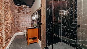 A bathroom with a brick wall, a wooden vanity, a black marble shower enclosure, and a tiled floor.