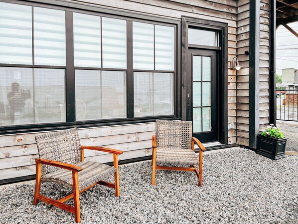 Zwei Korbstühle mit Holzgestellen auf einer Schotterterrasse neben einem Gebäude mit großen Fenstern und einer Tür.