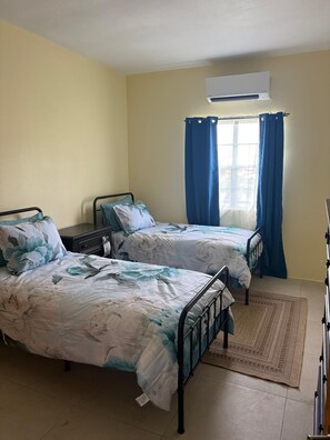 A room with two beds, a window with blue curtains, an air conditioning unit, and a woven rug on the floor. - Manna Heights. Perfect 2-bedroom apartment with AC in peaceful Bolands. (Bolands)
