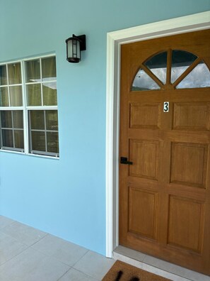 A wooden door with a number plate, a window with multiple panes, and a wall-mounted light fixture. - Manna Heights. Perfect 2-bedroom apartment with AC in peaceful Bolands. (Bolands)
