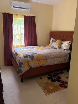 A bedroom with a bed, curtains, an air conditioner, and a floral rug. - Manna Heights. Perfect 2-bedroom apartment with AC in peaceful Bolands. (Bolands)