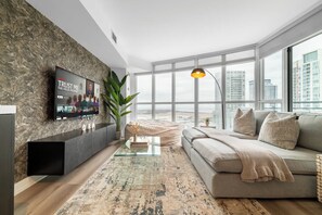 A modern living room with a large flat-screen TV, a sofa, a coffee table, and a large window offering a city view. - Waterfront 2BR Suite, Luxury Views & Parking | Parking Available (Toronto)