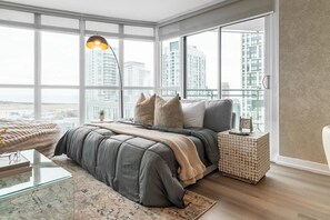 A modern bedroom with a large bed, a side table, and a view of a cityscape through large windows. - Waterfront 2BR Suite, Luxury Views & Parking | Parking Available (Toronto)