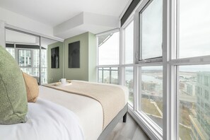 A modern hotel room with a large bed, a view of the cityscape, and a city skyline visible through the windows. - Waterfront 2BR Suite, Luxury Views & Parking | Parking Available (Toronto)