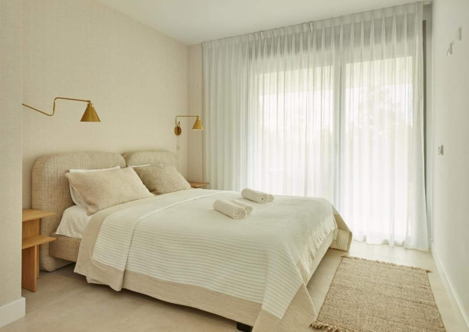 Een slaapkamer met een bed, twee bedlampen, een raam met dunne gordijnen en een geweven vloerkleed.