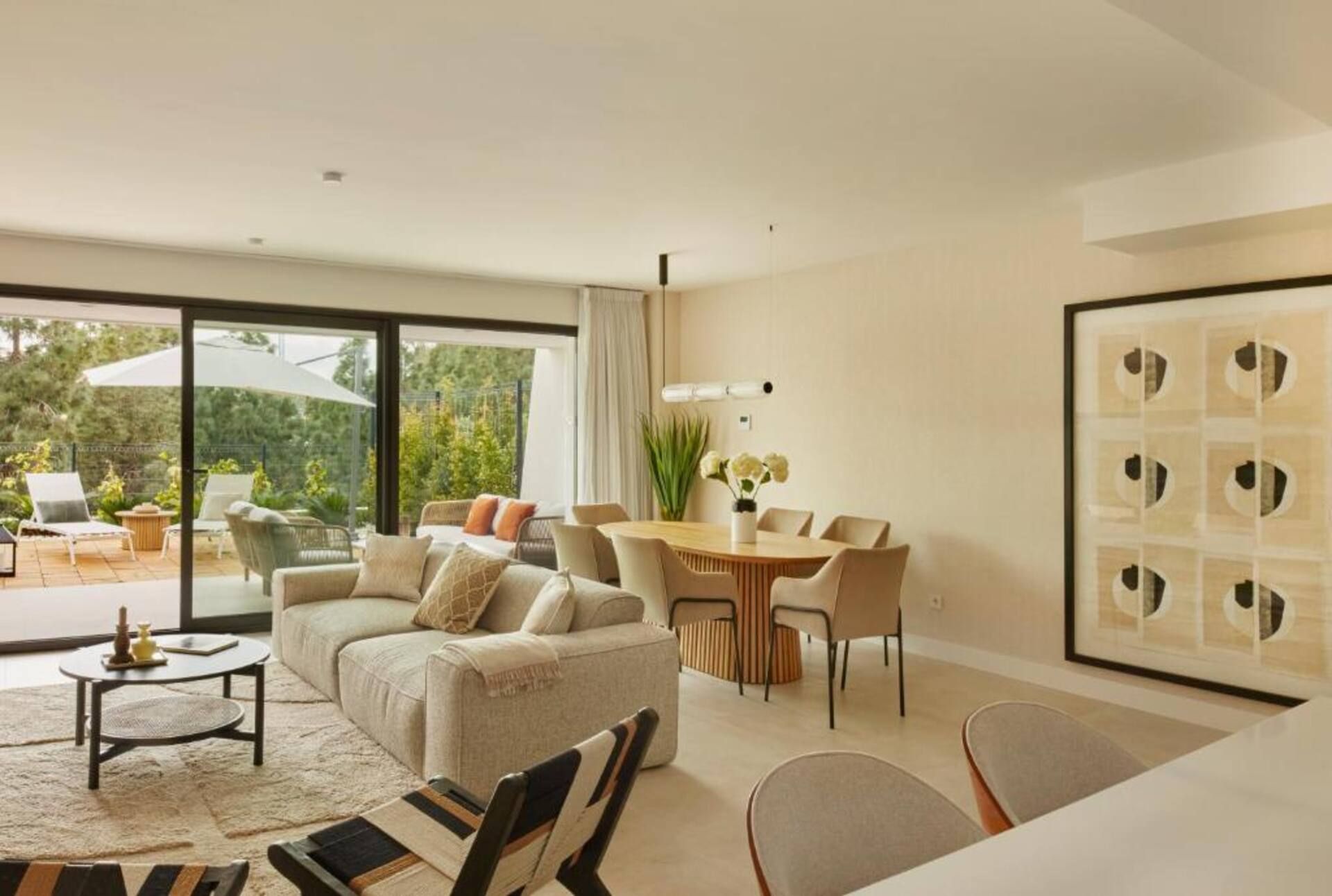 A modern living room with a sofa, coffee table, and dining table. There is a large window with a view of greenery.