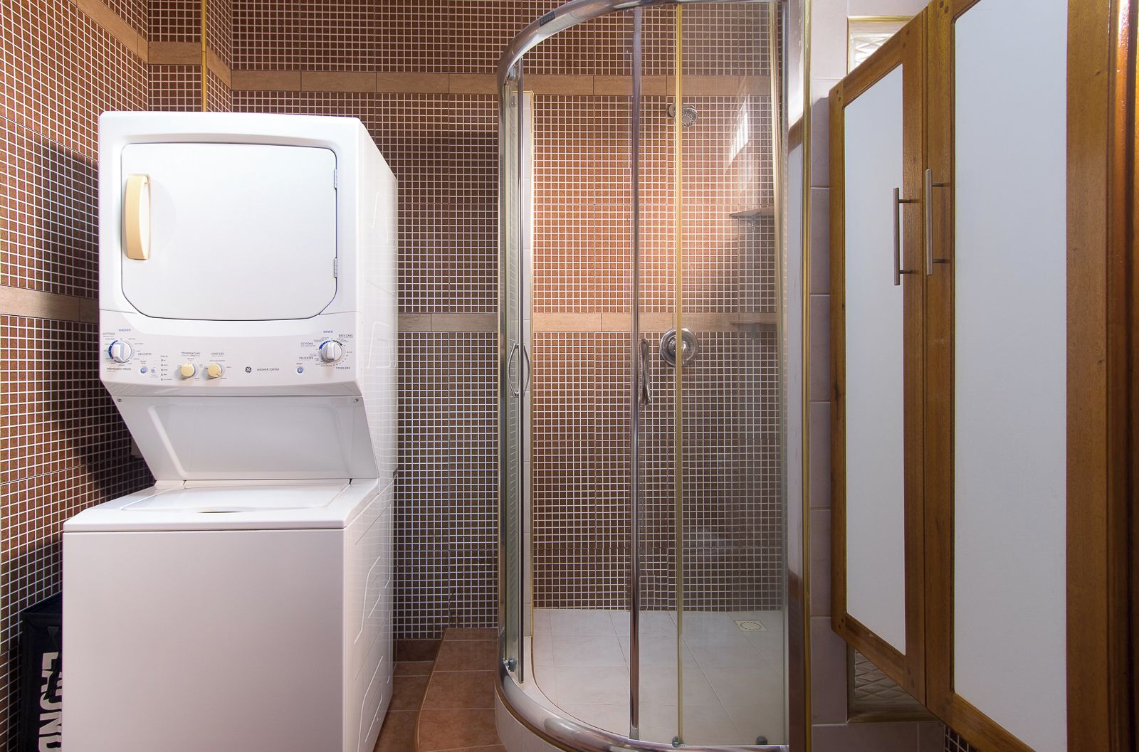 A compact bathroom with a washing machine, a shower enclosure, and a wooden cabinet.