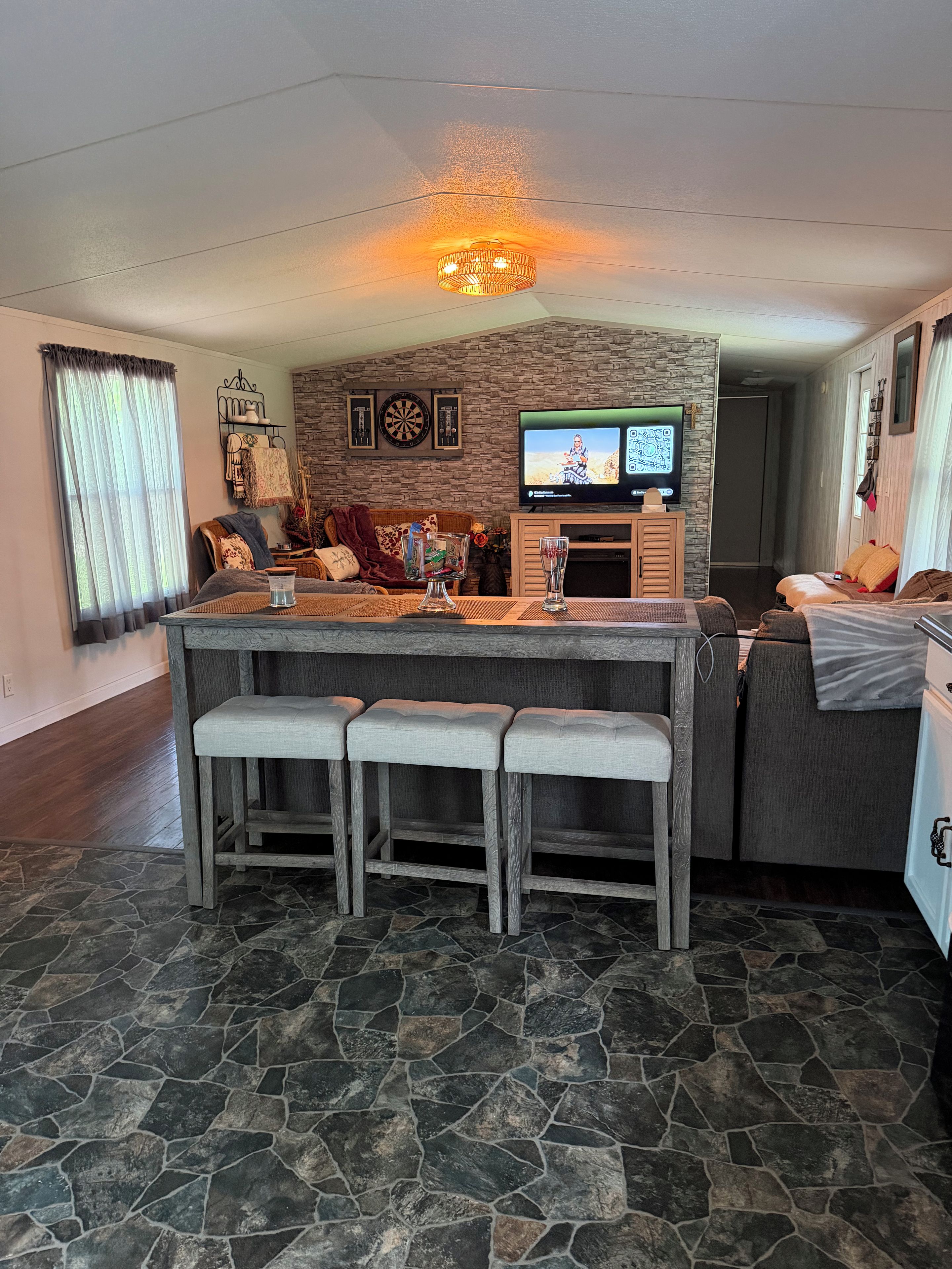A modern living room with a stone floor, a central island with bar stools, a flat-screen TV, and a fireplace.