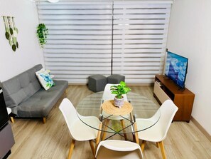 A modern living room with a grey sofa, a small round table with a plant, two white chairs, a TV on a wooden stand, and a window with blinds.