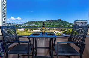 A balcony with two chairs and a table, overlooking a cityscape with a mountain in the distance. - [New] Modern Luxe 2 Bedroom Condo in Waikiki with Diamond Head Views! (Honolulu)