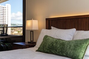 Habitación de hotel con cama, mesita de noche, lámpara y ventana con vistas a edificios.