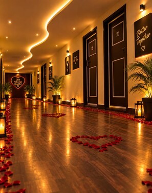 A hotel corridor with a wooden floor, red rose petals, wall-mounted picture frames, and a heart-shaped sign with the text 'Love Makes Life Beautiful.' - Hotel East Park Rio (Anekal)