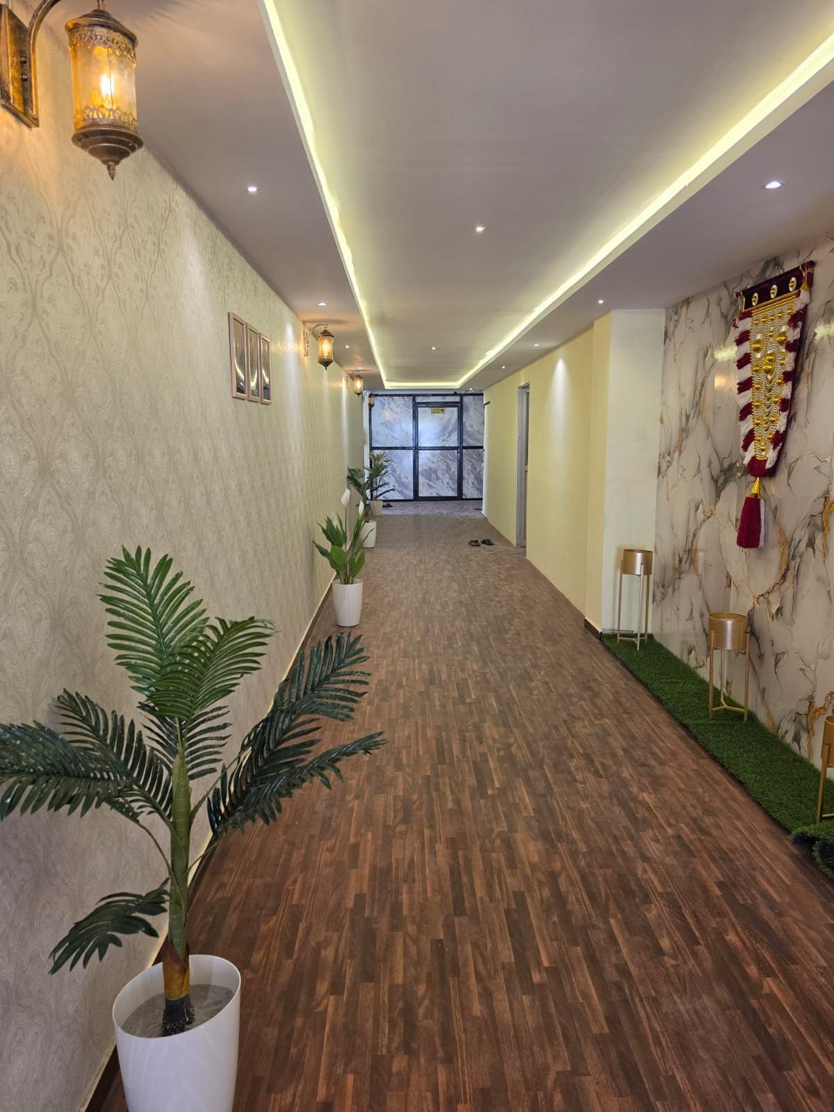 A hotel lobby with a wooden floor, potted plants, wall-mounted lights, and a marble wall with a decorative banner.