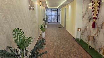 Un hall de hotel con piso de madera, plantas en macetas, luces fijadas a la pared y una pared de mármol con un estandarte decorativo.