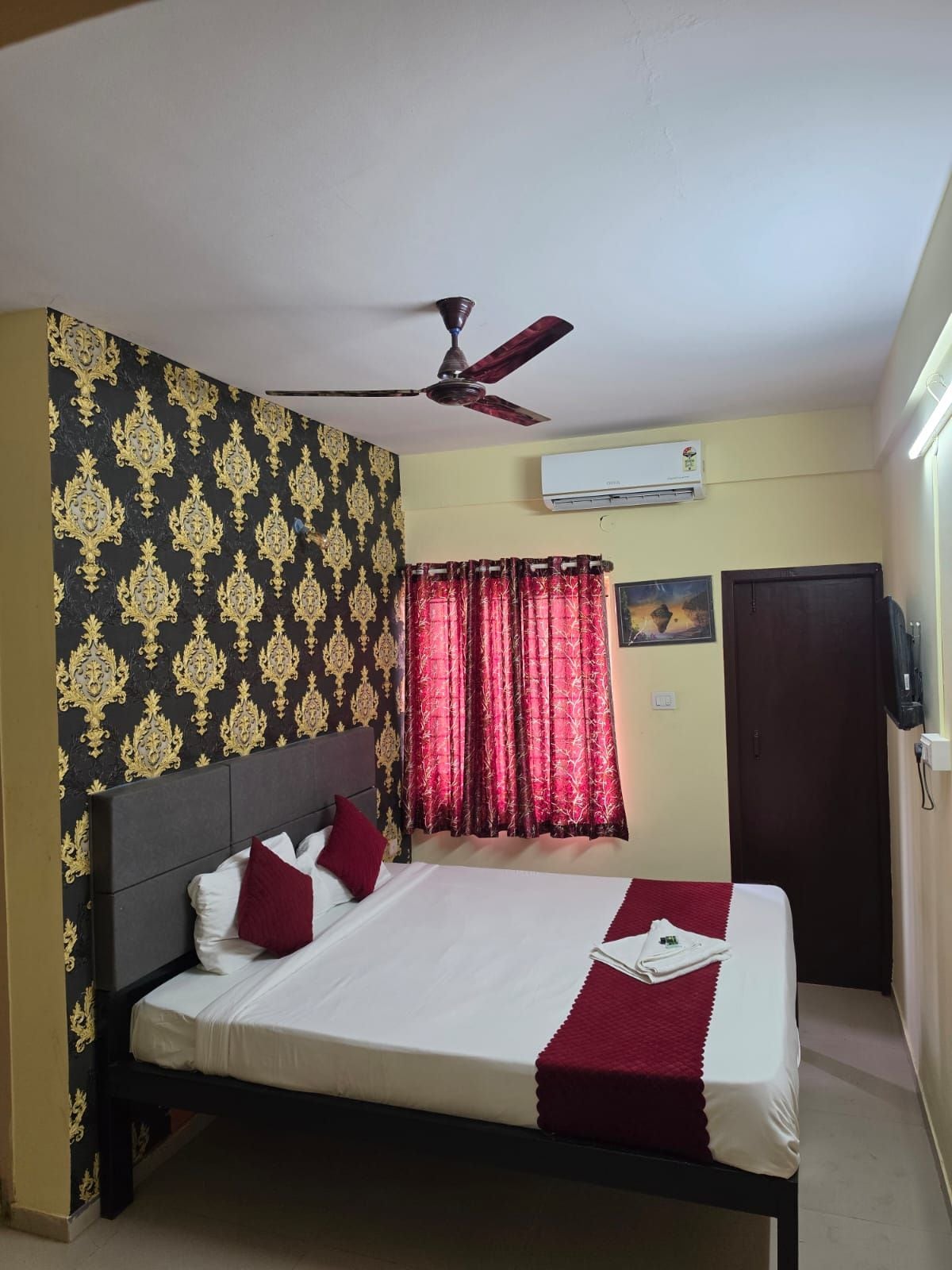 Habitación de hotel con cama, ventilador de techo, equipo de aire acondicionado, una puerta y una ventana con cortinas.