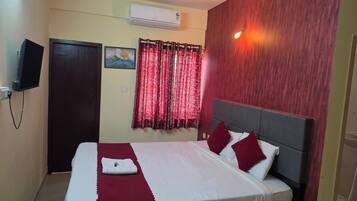 Habitación de hotel con cama, televisor de pantalla plana en la pared, equipo de aire acondicionado y ventana con cortinas rojas.