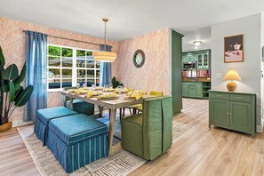 Ein Essbereich mit einem Tisch für vier Personen, einem grünen Sofa, einem großen Fenster mit Vorhängen und einer Küche im Hintergrund.