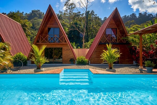 A pool area with three wooden triangular cabins, each with a balcony, surrounded by tropical plants. - SINA BUNGALOWS PUERTO VIEJO (Cahuita)