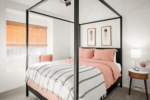 A four-poster bed with a striped bedspread and pink pillows, a bedside table with a lamp, and framed artwork on the wall. - Lakefront Glendale Oasis - Private Pool & Hot Tub (Glendale)
