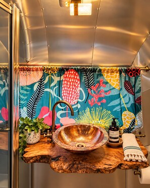 A bathroom with a wooden vanity, a copper sink, a potted plant, and a colorful shower curtain. - Romantic Vintage Airstream Safari Near Puget Sound (Des Moines)