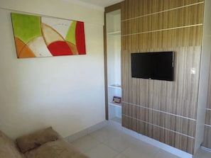 Una habitación con un televisor de pantalla plana montado en una pared de paneles de madera, un cuadro abstracto y colorido en la pared, y un sofá blanco con un cojín.