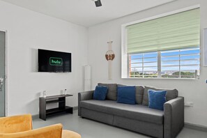 A modern living room with a grey sofa, a flat-screen TV, a window with blinds, and a yellow chair. - Santurce Blue 2BR | Near Beach & La Placita (San)
