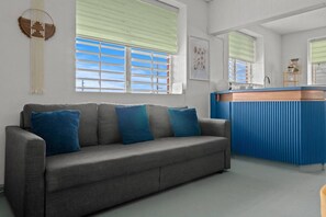 Ein graues Sofa mit blauen Kissen, eine blaue Küchenzeile und ein Fenster mit Jalousien.