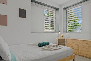 Ein Schlafzimmer mit einem Bett, einer hölzernen Kommode, zwei Fenstern mit Jalousien und einer Wandleuchte.