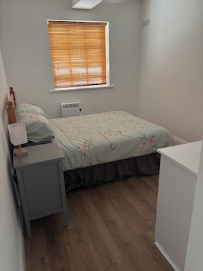A bedroom with a bed, a bedside table with a lamp, a radiator, and a window with blinds.