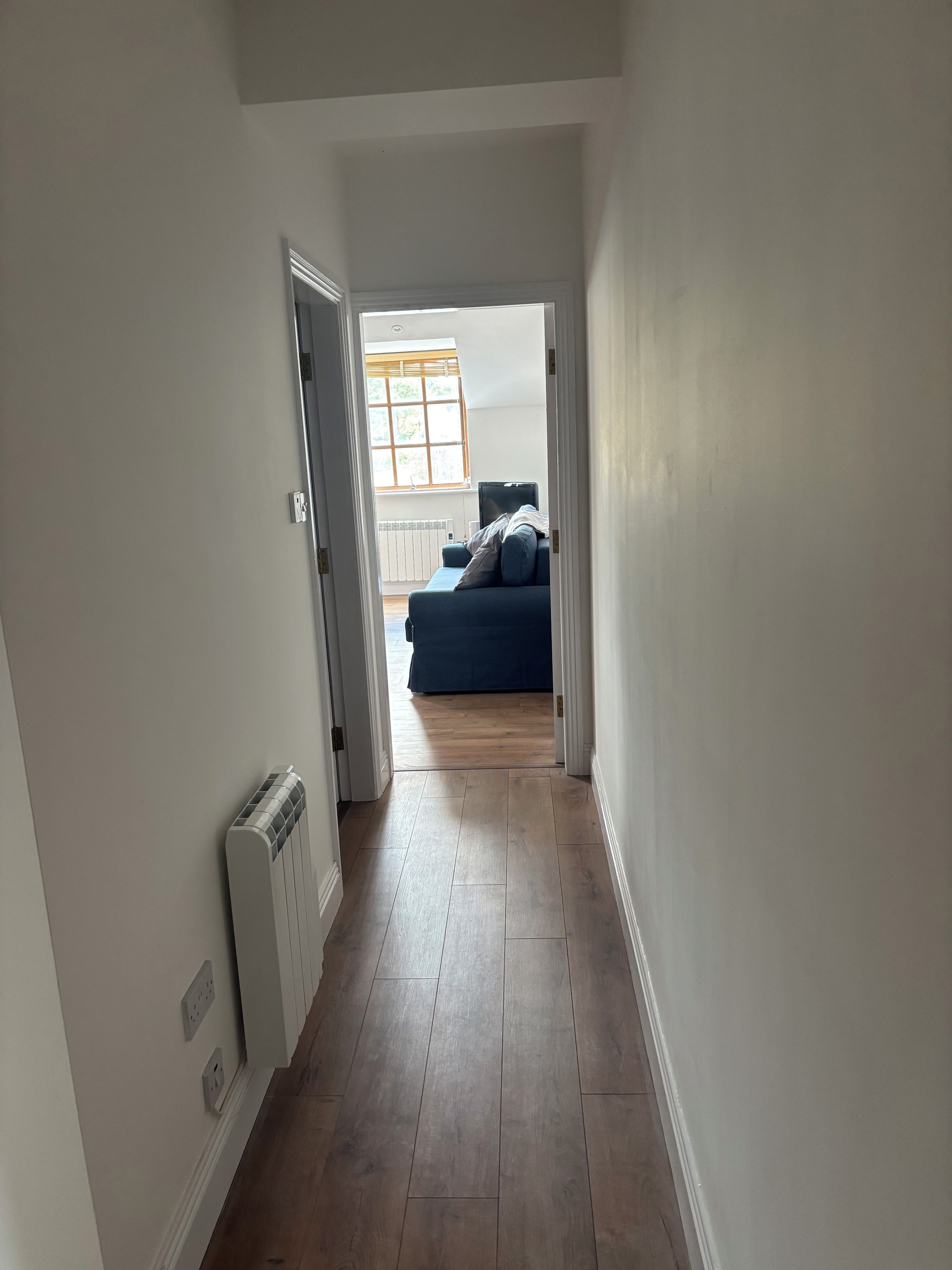 A hallway with a radiator, a window with blinds, and a blue sofa in the distance.
