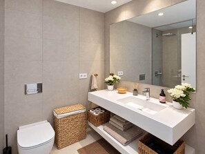 Een moderne badkamer met een witte wastafel, spiegel en toilet. Er staan rieten manden en een kleine plant op het aanrecht.
