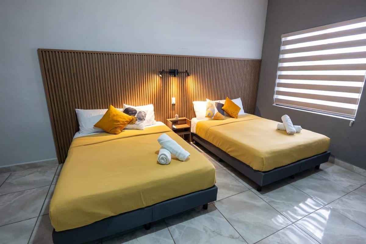 Habitación de hotel con dos camas, cada una con cubrecama amarillo, cabecera de madera y mesita de noche con lámpara.