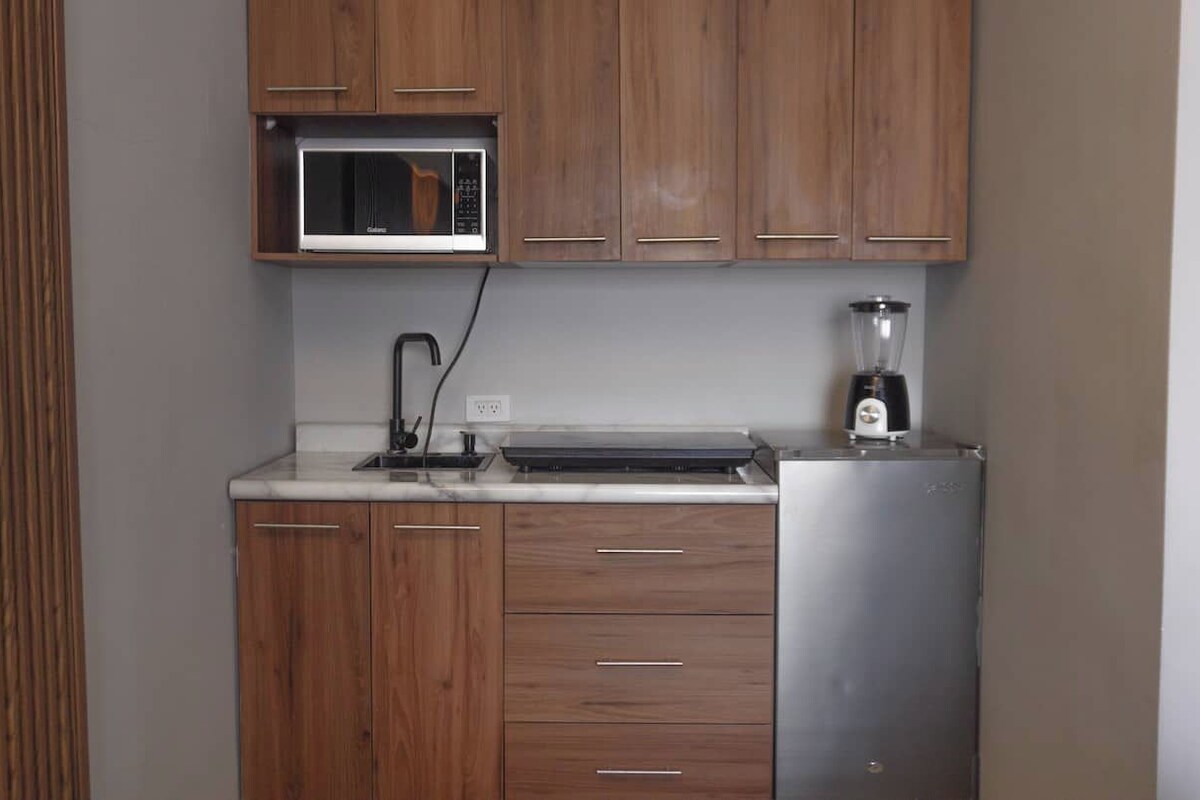 A compact kitchen with wooden cabinets, a built-in microwave, a blender, and a refrigerator.