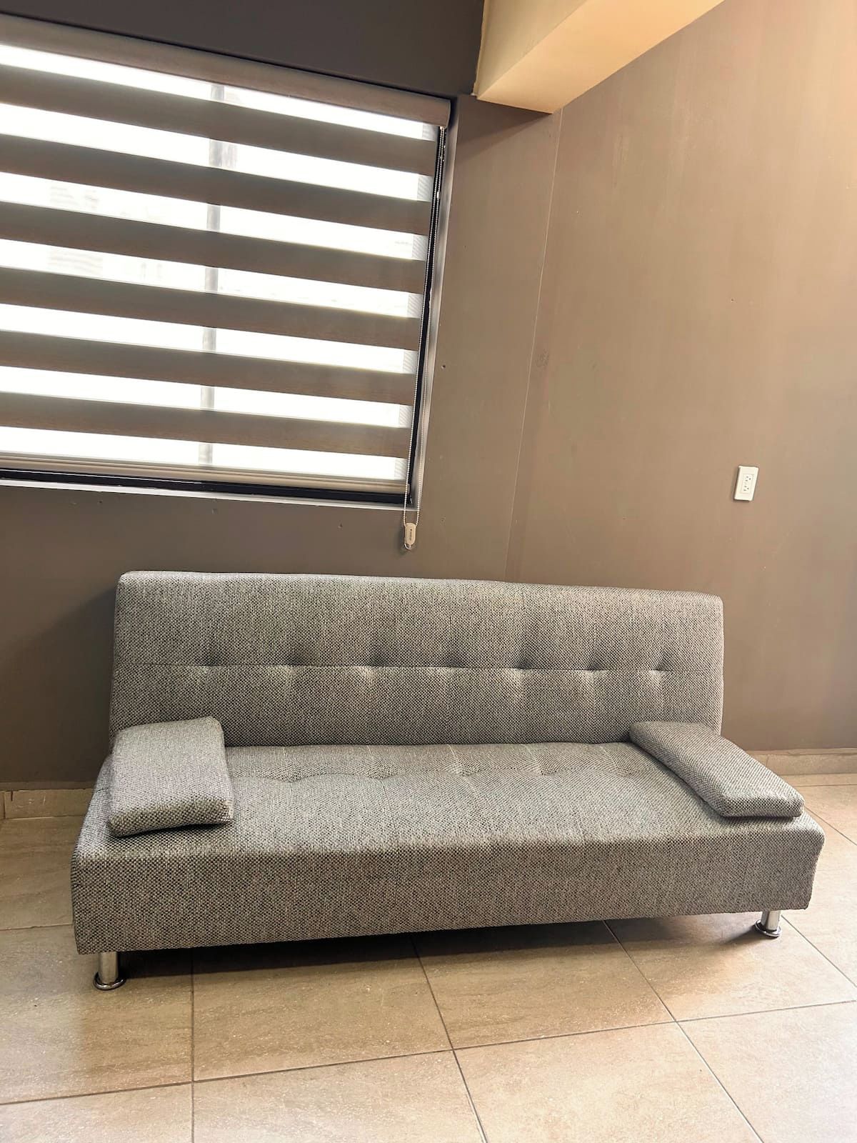 A grey sofa with a matching pillow, positioned in front of a window with horizontal blinds.