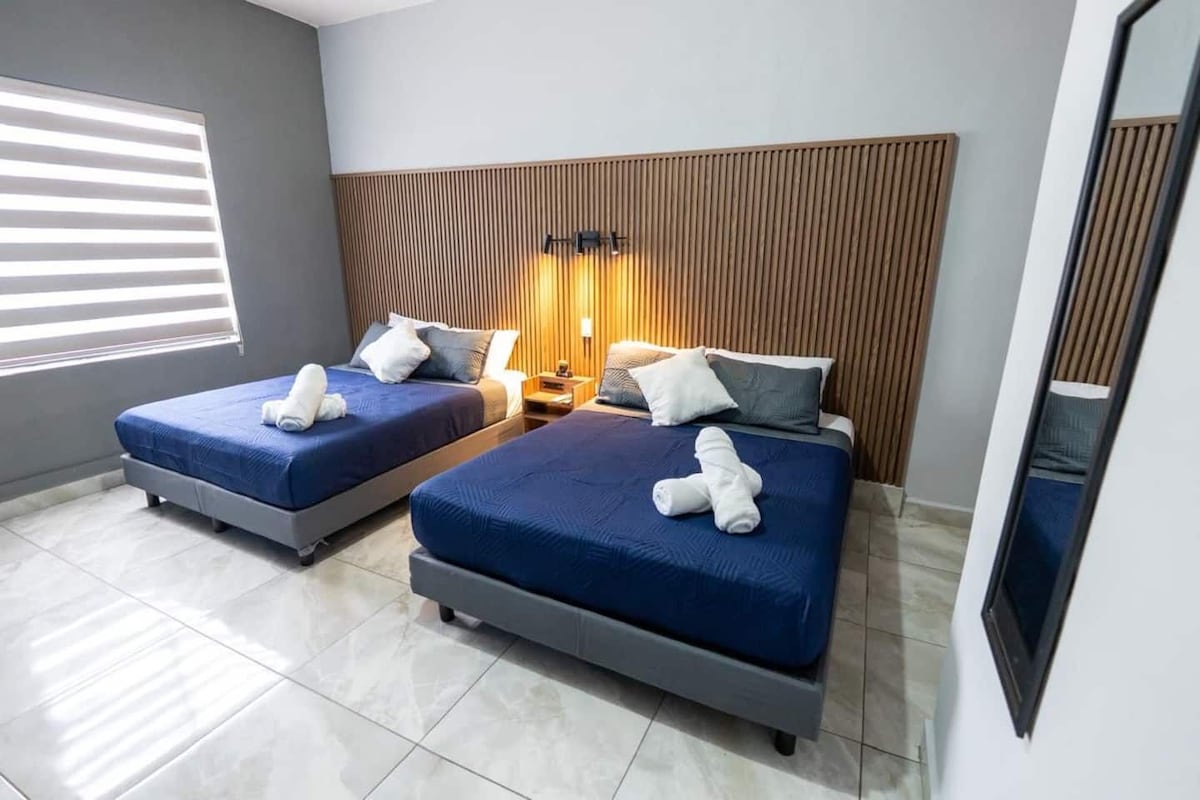 Quarto de hotel com duas camas, cabeceira de madeira, mesa de cabeceira com abajur e um espelho grande.