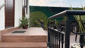 A set of wooden steps leading to a door, a metal railing, and a green wall with a hedge.