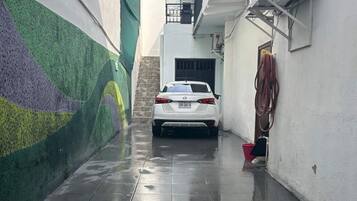 A narrow alleyway with a white car parked in the center, a green wall on the left, and an air conditioning unit on the right.