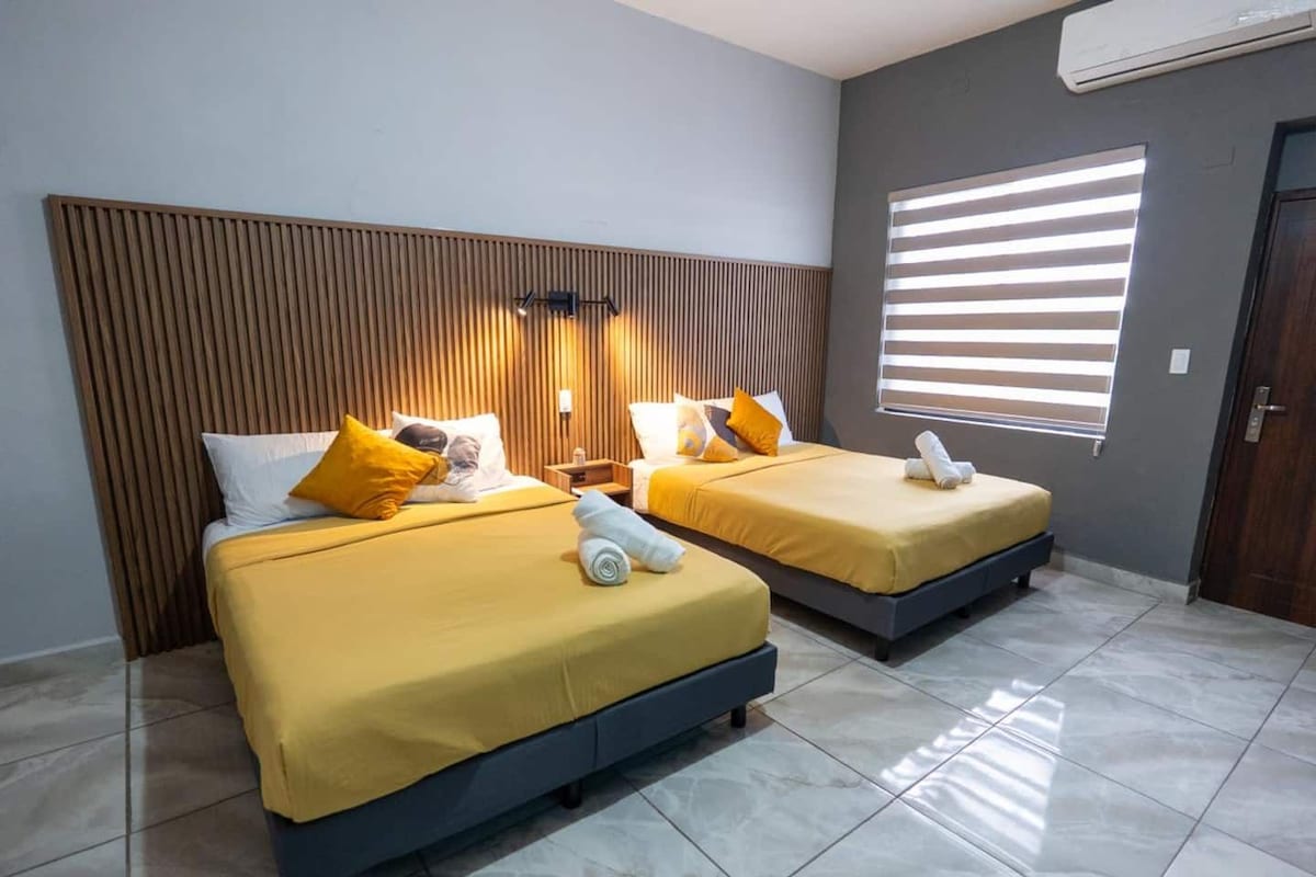 Quarto de hotel com duas camas, cabeceira de madeira, janela com persianas e piso de azulejos.
