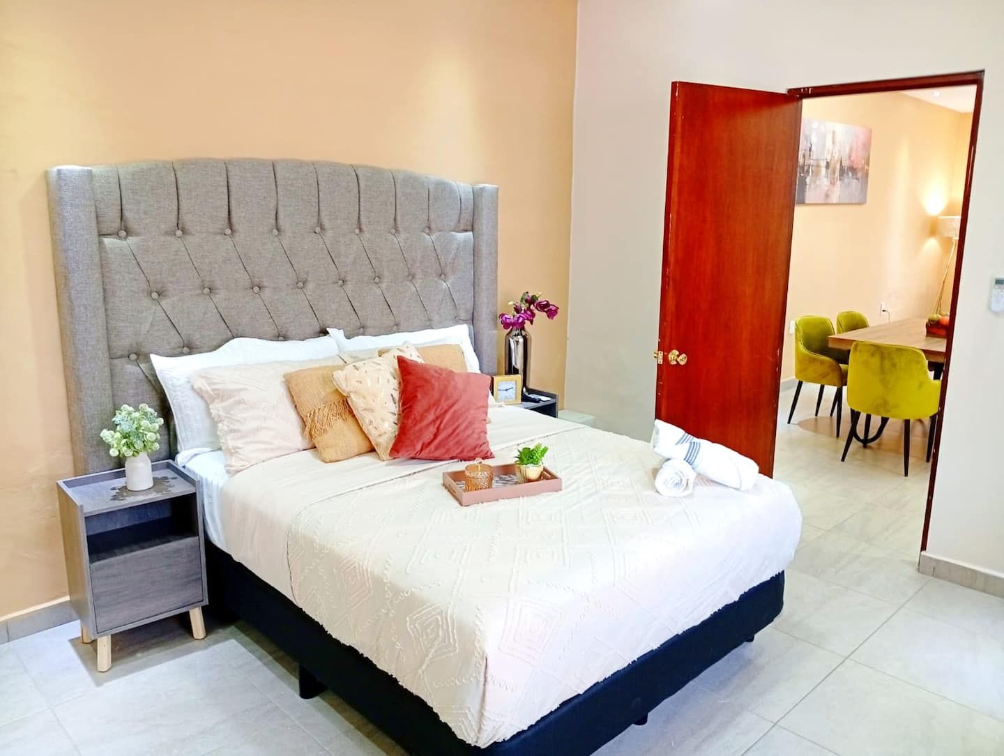 Una cama bien tendida con un cabecero tapizado en gris, una mesita de noche con un florero y vistas hacia un comedor con sillas amarillas.