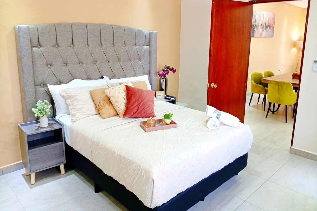 A neatly made bed with a grey upholstered headboard, a bedside table with a vase of flowers, and a view into a dining area with yellow chairs.