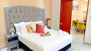 Una cama bien tendida con un cabecero tapizado en gris, una mesita de noche con un florero y vistas hacia un comedor con sillas amarillas.