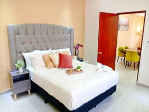 A neatly made bed with a grey upholstered headboard, a bedside table with a vase of flowers, and a view into a dining area with yellow chairs. - Casa Arteaga (Monterrey)