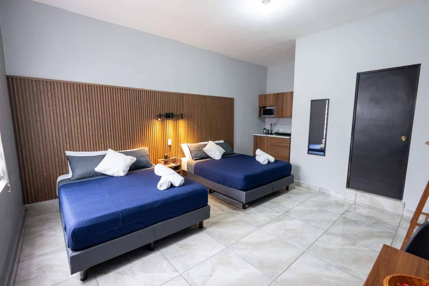 Habitación de hotel con dos camas, cabecera de madera, kitchenette pequeña y una puerta.